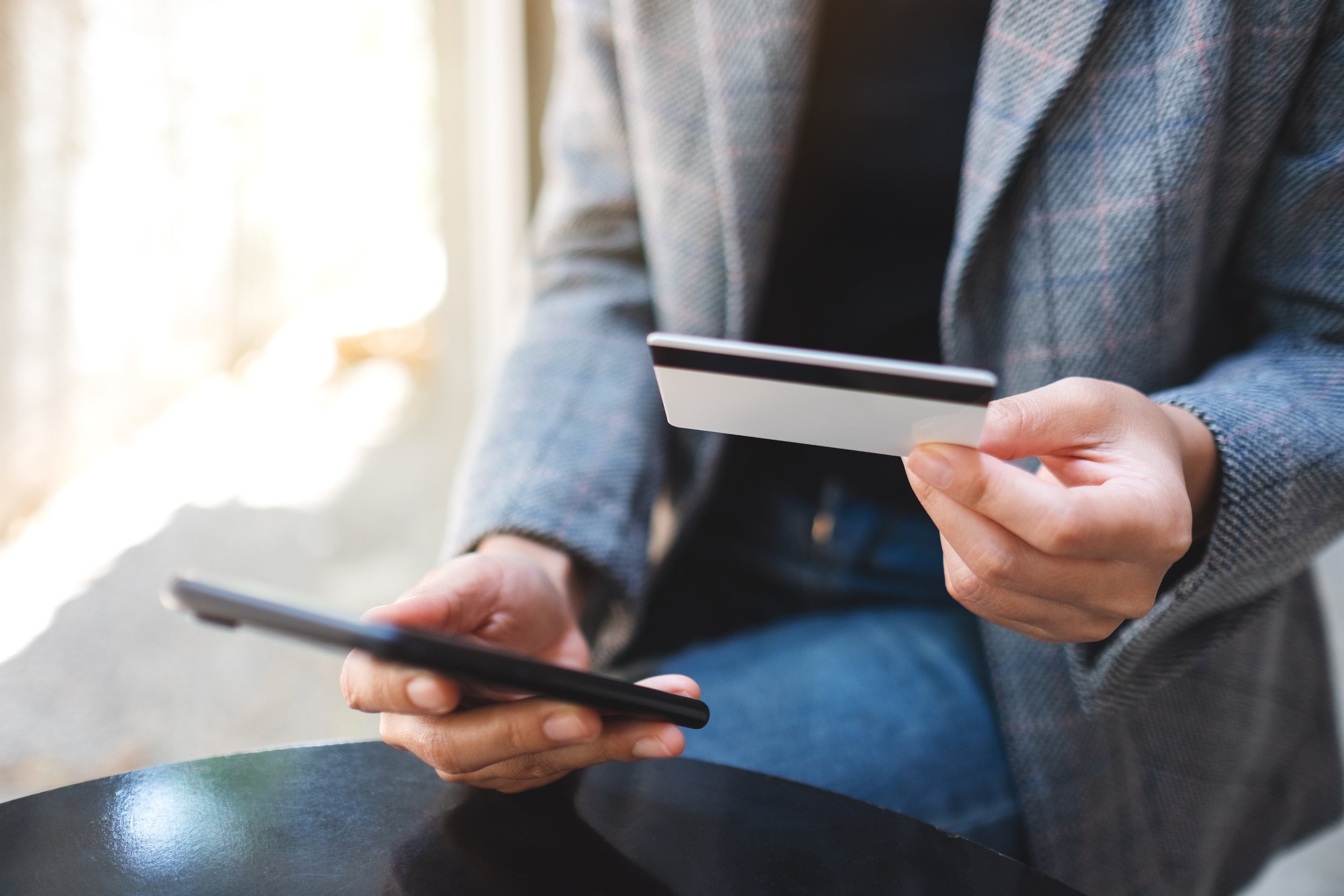 A person in a gray checkered blazer holding a credit card in one hand and a smartphone in the other, seated at a table.