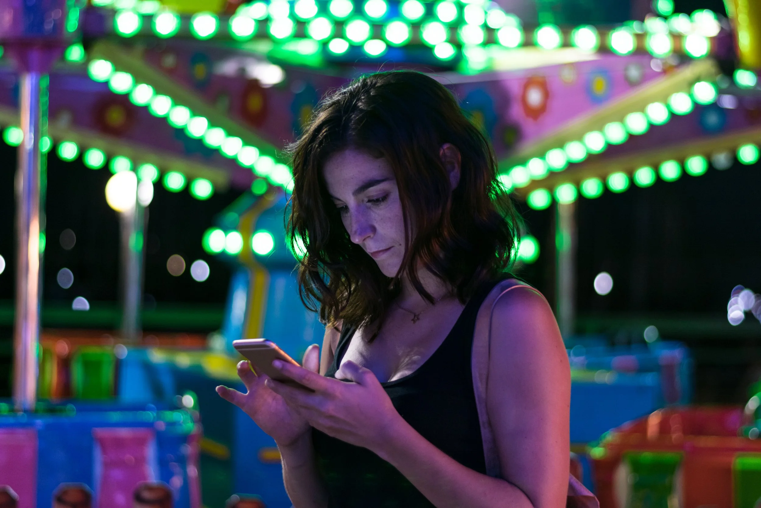 A young woman with dark, wavy hair looking at her phone at a nighttime amusement park with colorful, illuminated rides and lights in the background.