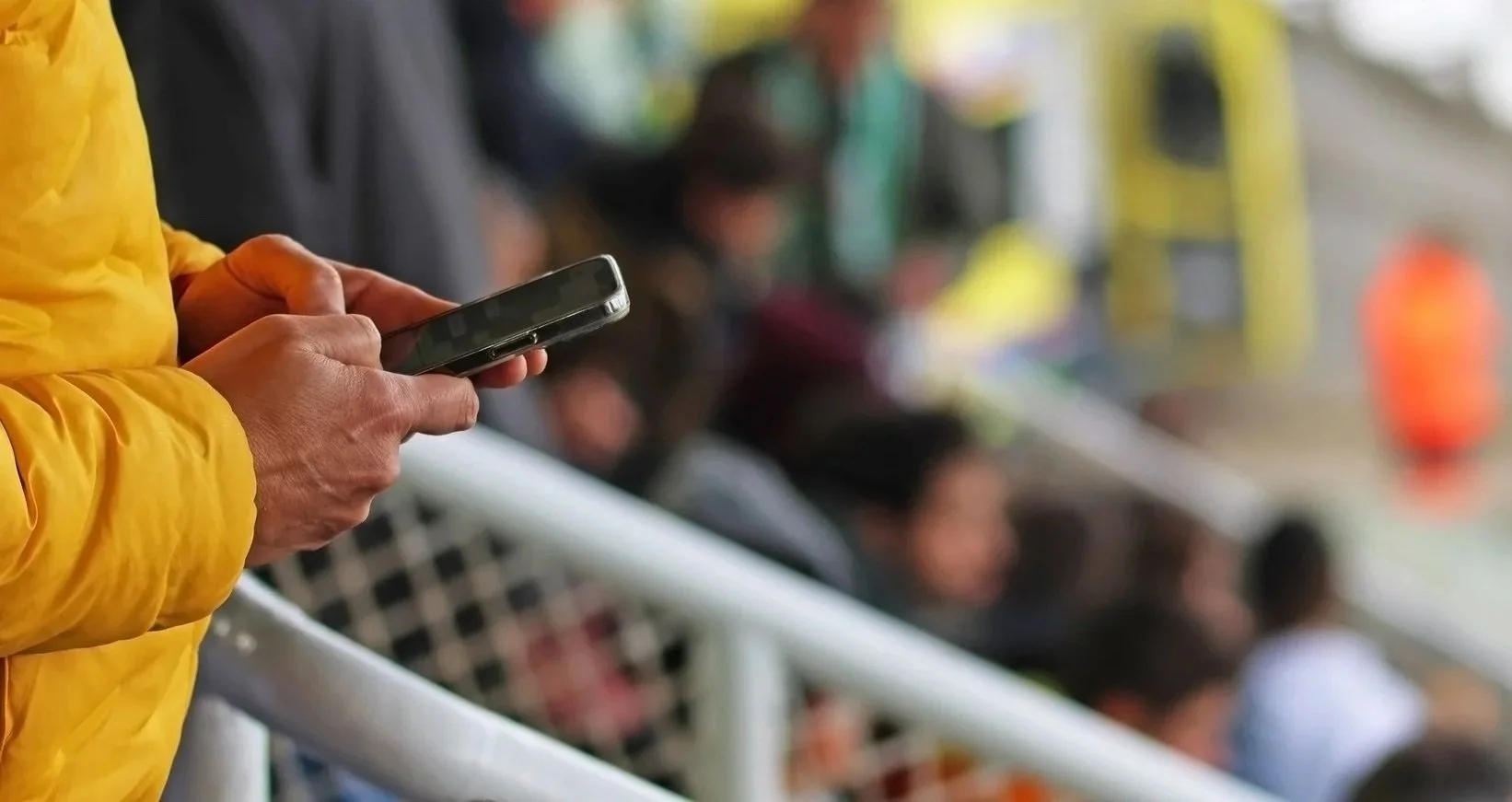 Person in a yellow jacket using a smartphone at a stadium or outdoor event with blurred seated crowd in the background.