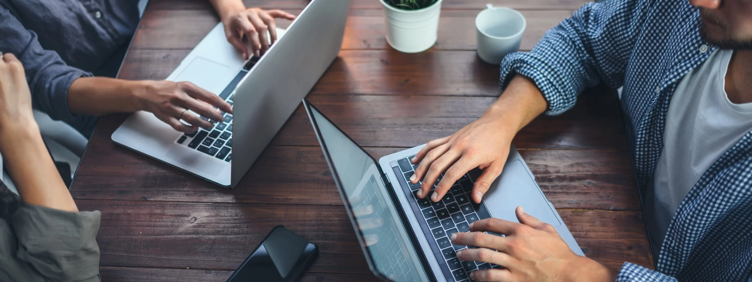 Group of people working on laptops at a wooden table, with some coffee mugs and a potted plant nearby.