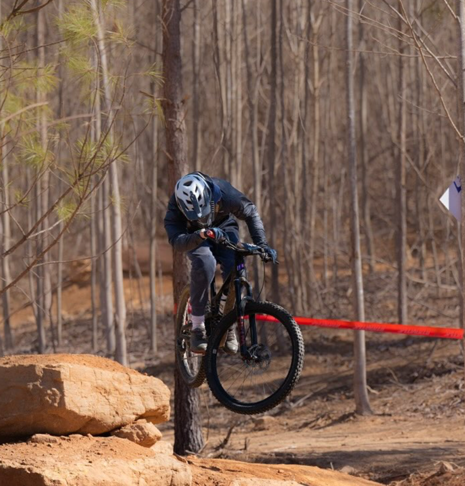 A person wearing a helmet and protective gear riding a mountain bike and jumping over a rock on a dirt trail in a wooded area.