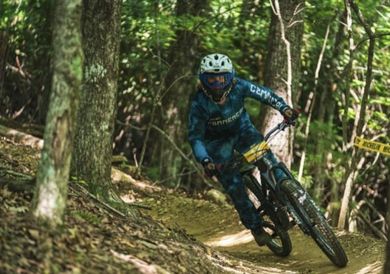 A person riding a mountain bike on a dirt trail in a forest, wearing a helmet, goggles, and a blue and black outfit.