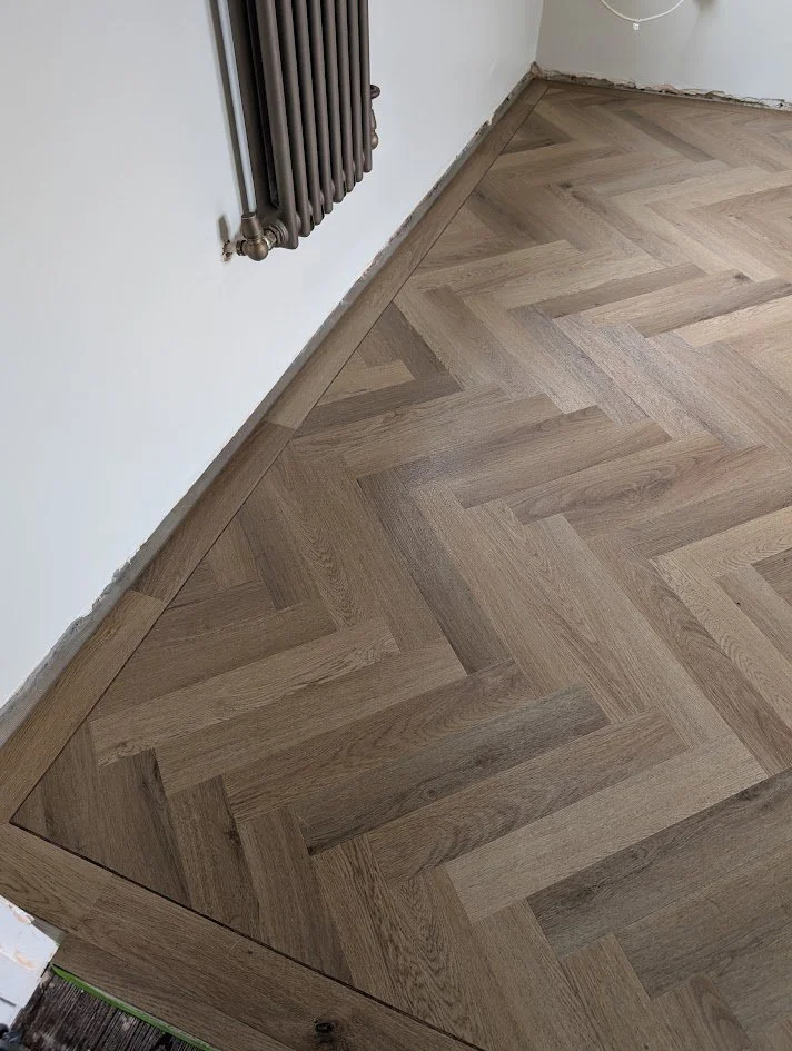 Close-up of a wooden herringbone parquet floor arranged in a herringbone pattern next to a white wall with a radiator attached.