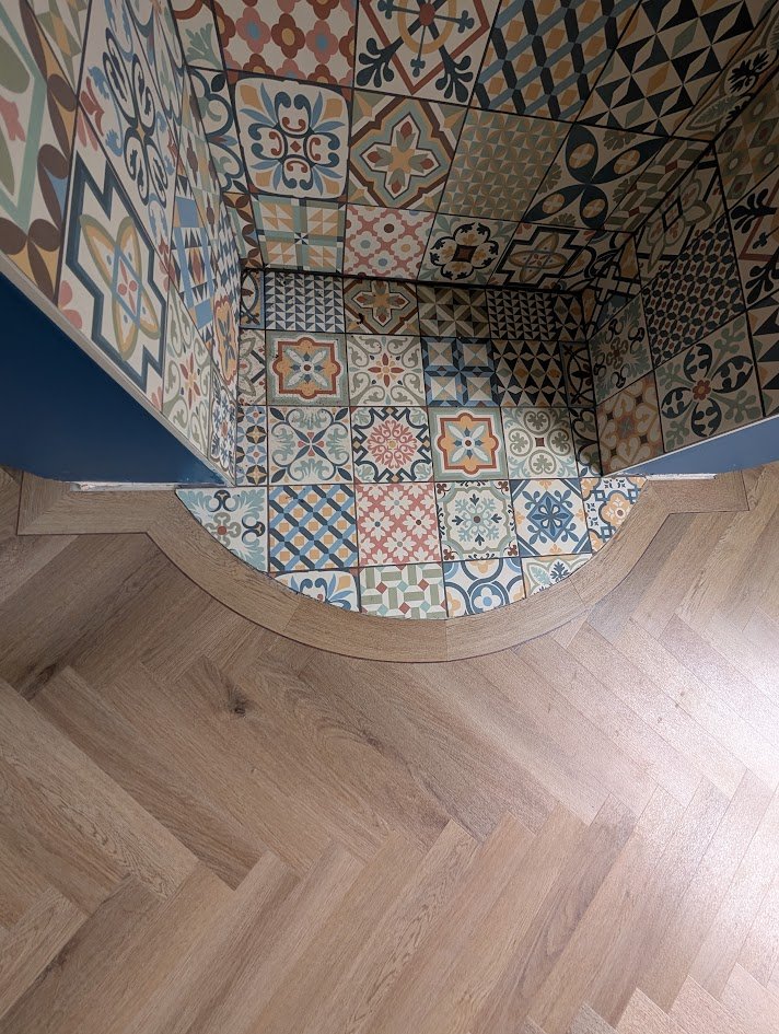 Part of a staircase with colorful patterned tiles on the side wall and floor, and wooden flooring at the bottom.