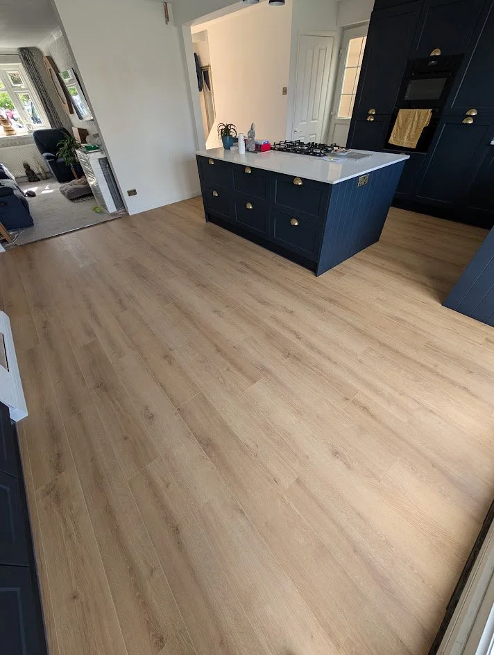 Empty kitchen with light wood flooring and navy blue cabinets, a kitchen island with a white countertop, and a gas cooktop. In the background, there is a doorway leading to a living room with large windows and some furniture.