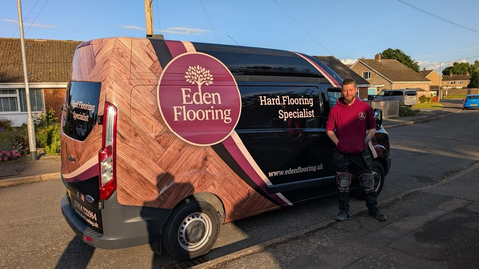 A man standing beside a black van with Eden Flooring branding and wood floor images, in a suburban neighborhood.