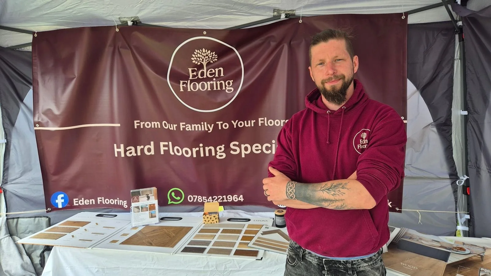 A man standing in front of a booth for Eden Flooring, wearing a maroon hoodie with the company logo. The booth has samples of flooring and a large banner displaying the Eden Flooring logo, contact information, and the slogan "From Our Family To Your Floor".