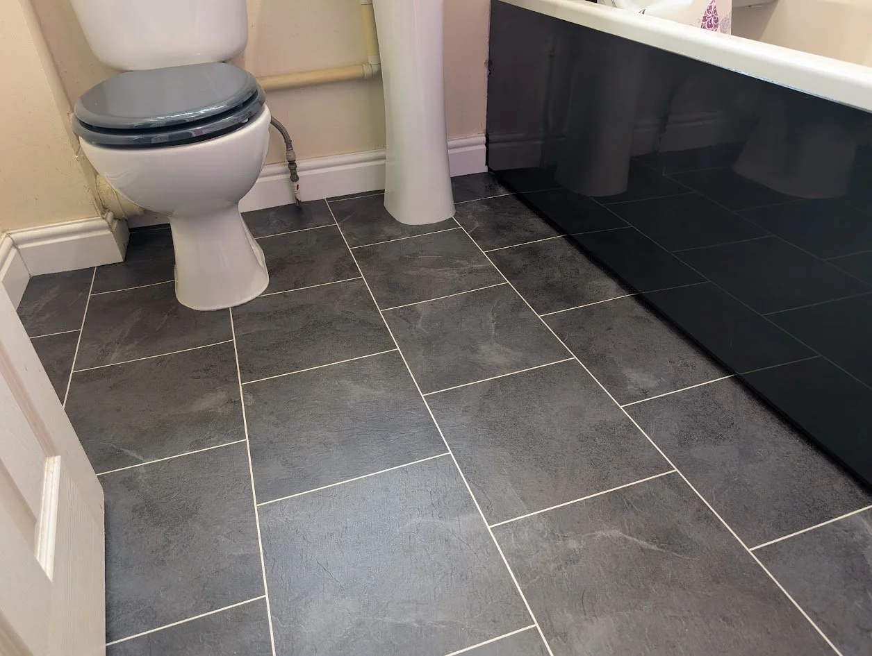 Bathroom with a white toilet with a gray seat, a white sink, and black glossy cabinet, all on black LVT tiles with white grout strips.