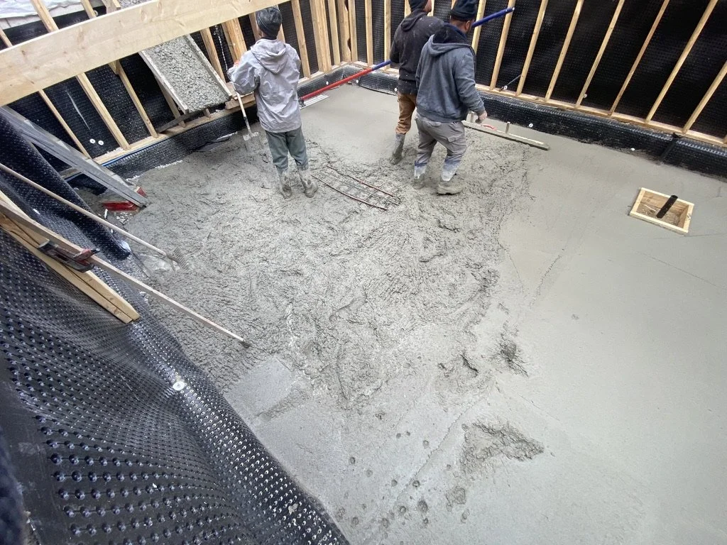 Workers pouring and leveling concrete on a construction site, with wooden framing and black waterproofing membrane visible around the floor.