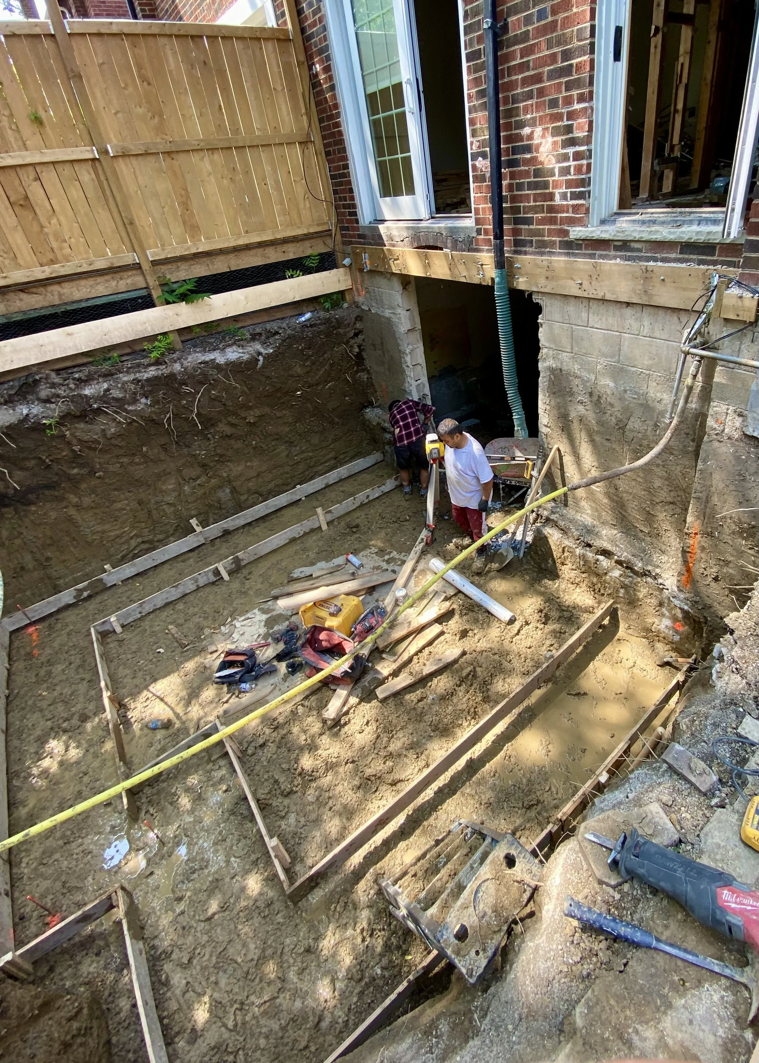 Construction workers excavating a backyard to prepare for building a foundation or basement, with tools and building materials scattered around.