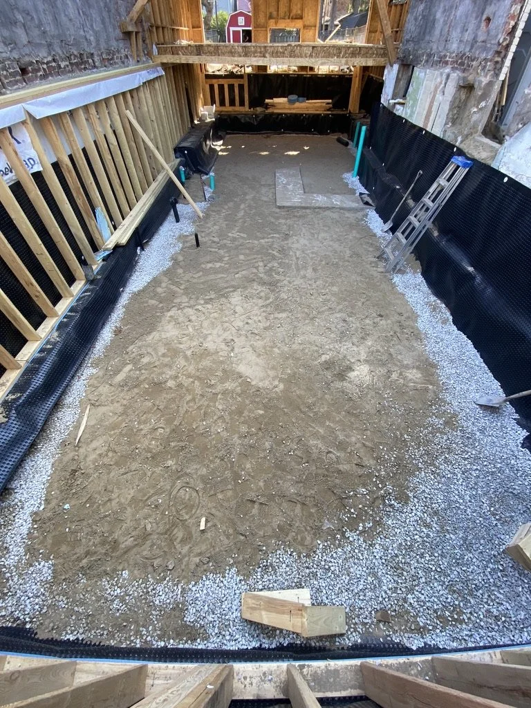 Construction site with a dirt floor, wooden and black drainage or vapor barrier lining, and framing for a new structure. A ladder rests against the right wall, and various tools and materials are visible along the edges.