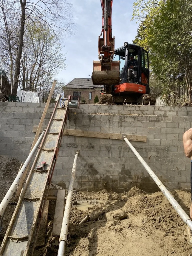 Construction worker operating a small excavator on a residential wall, with a makeshift ladder and support beams in a backyard.