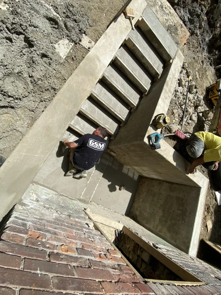 Construction workers installing concrete stairs on a building site with soil and brick wall nearby.