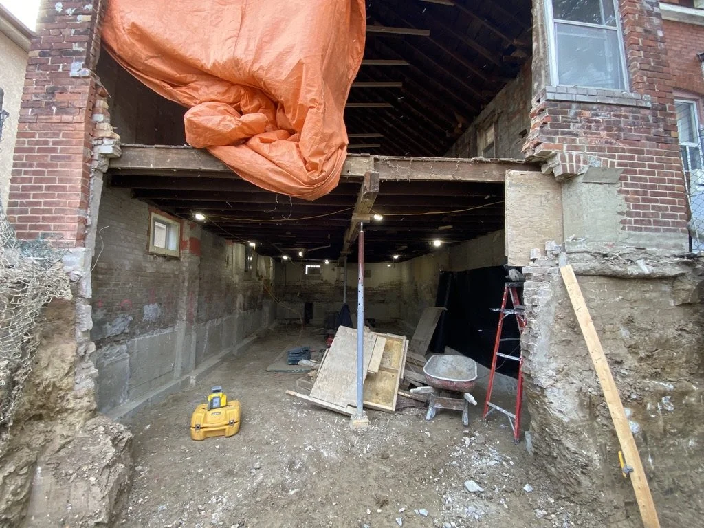 Construction site of a house renovation showing exposed brick walls, a partially removed floor, construction tools, and an orange tarp hanging from the ceiling.