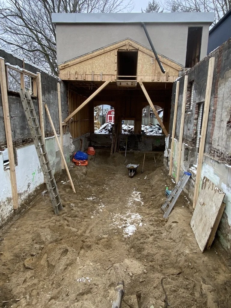 Construction site showing the interior of a house under renovation with exposed walls, ladders, and construction tools. The house has a new second floor or roof structure being built.
