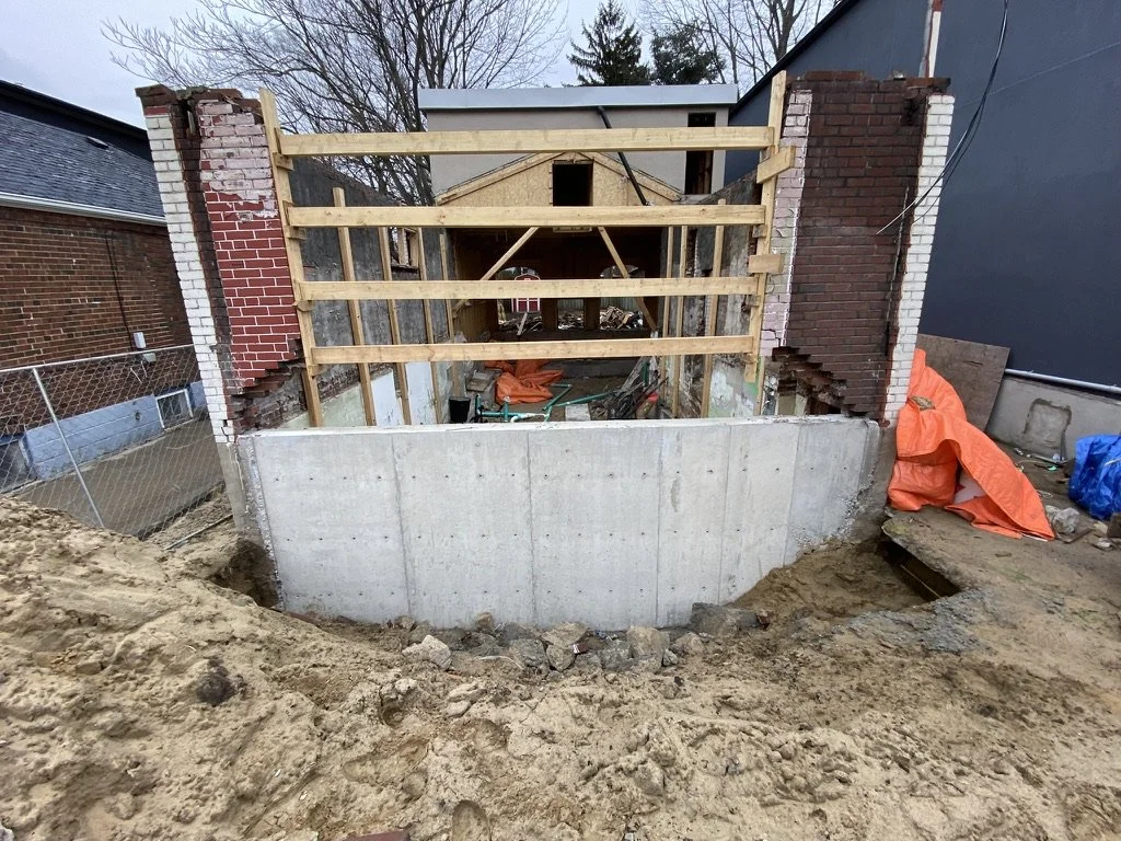 Construction site showing a partially built brick wall and wooden framing, with a concrete foundation and surrounding earth.