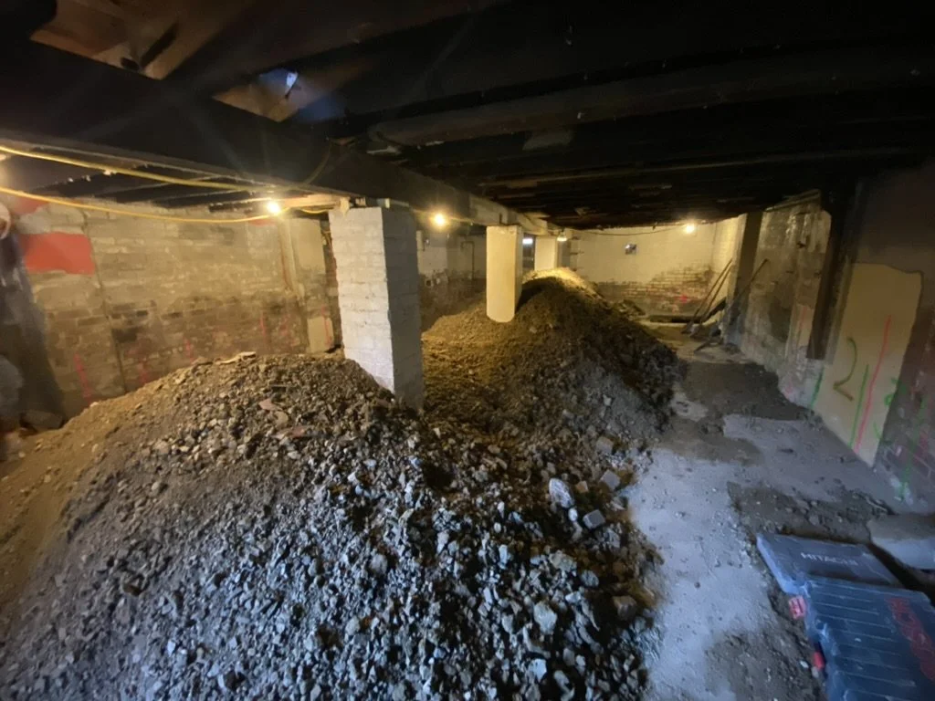 Construction site with piles of dirt and rubble under a ceiling with exposed pipes and construction lights.