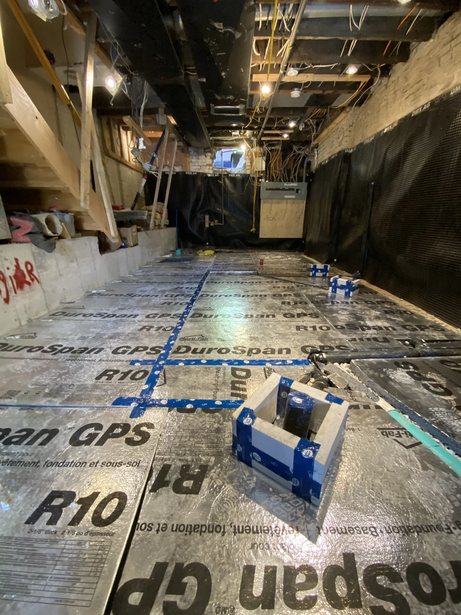 Interior of a building under construction with insulation, electrical wiring, and framing visible, showing a floor with waterproofing membrane and some blue electrical conduit.