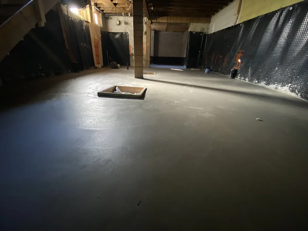 Unfinished attic with a freshly poured concrete floor, a wooden beam in the center, and insulation panels along the walls.