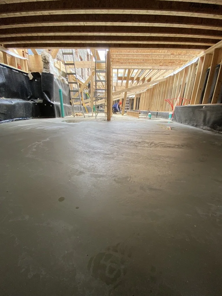 Construction site with a poured concrete floor, wooden framing, and a worker in the background.