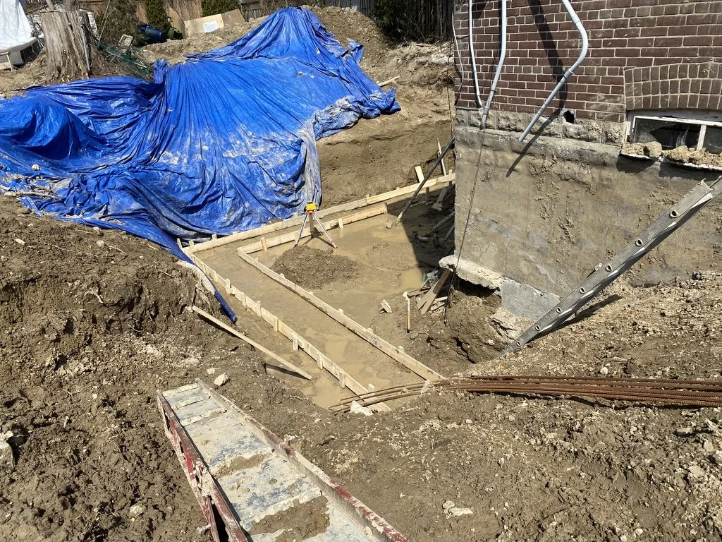 Construction site with a partially built foundation and a blue tarp covering part of the area, next to a brick wall.