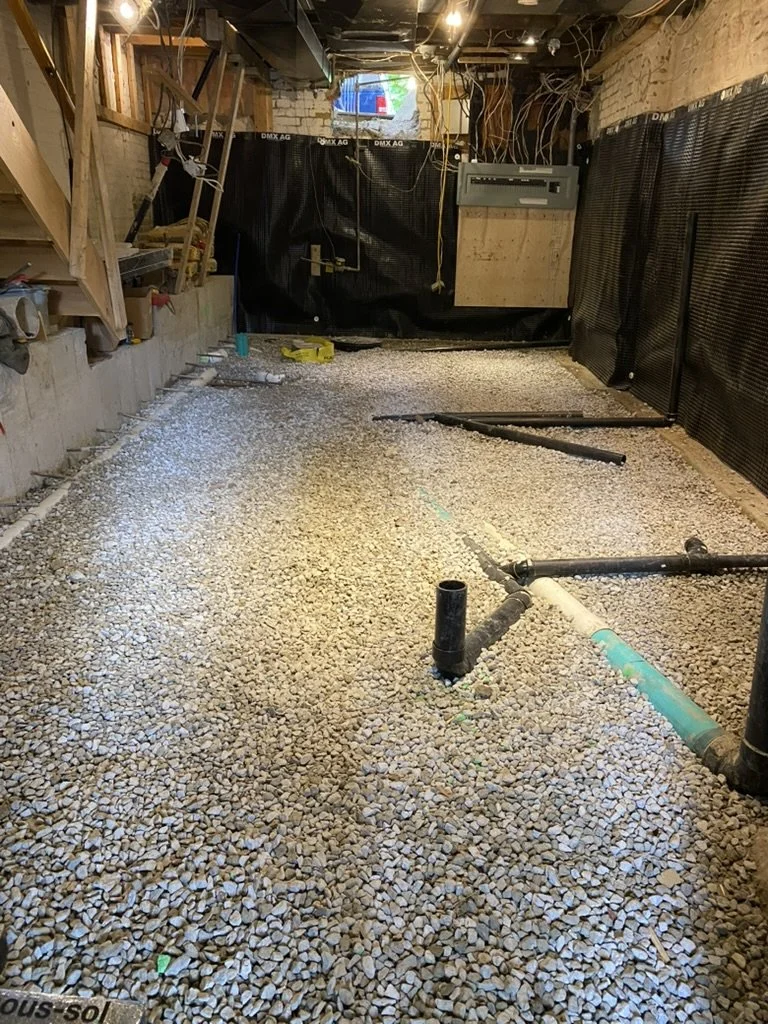 Interior of a construction site showing a gravel floor with exposed pipes, electrical wiring, and unfinished walls.