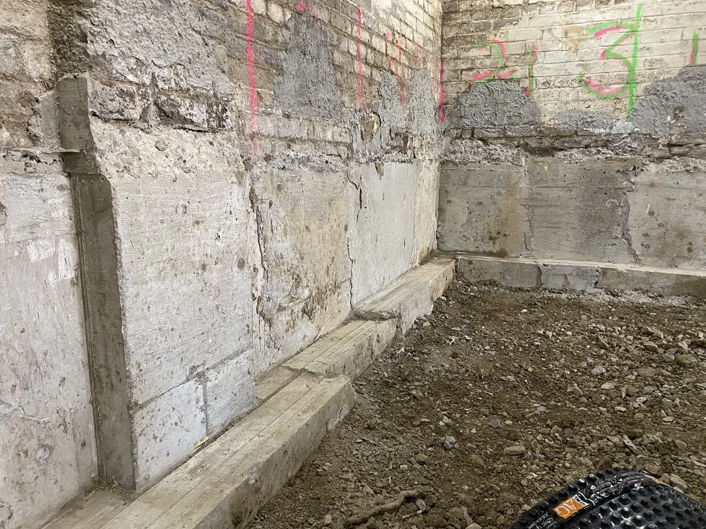 Interior view of a construction site with exposed brick walls, wooden beams along the base, and pink chalk markings and numbers on the brick wall.