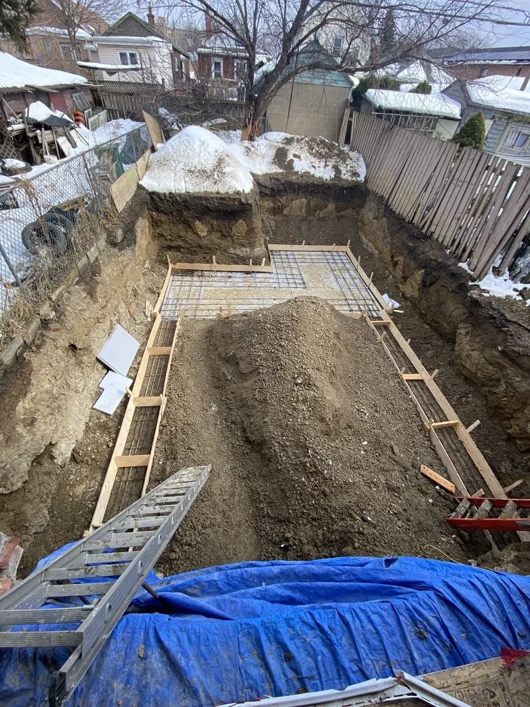 Construction site in a backyard, with an excavated area for a new foundation, surrounded by wooden forms, rebar grid, a ladder, and a pile of dirt. Snow is visible on the ground and on rocks outdoors.