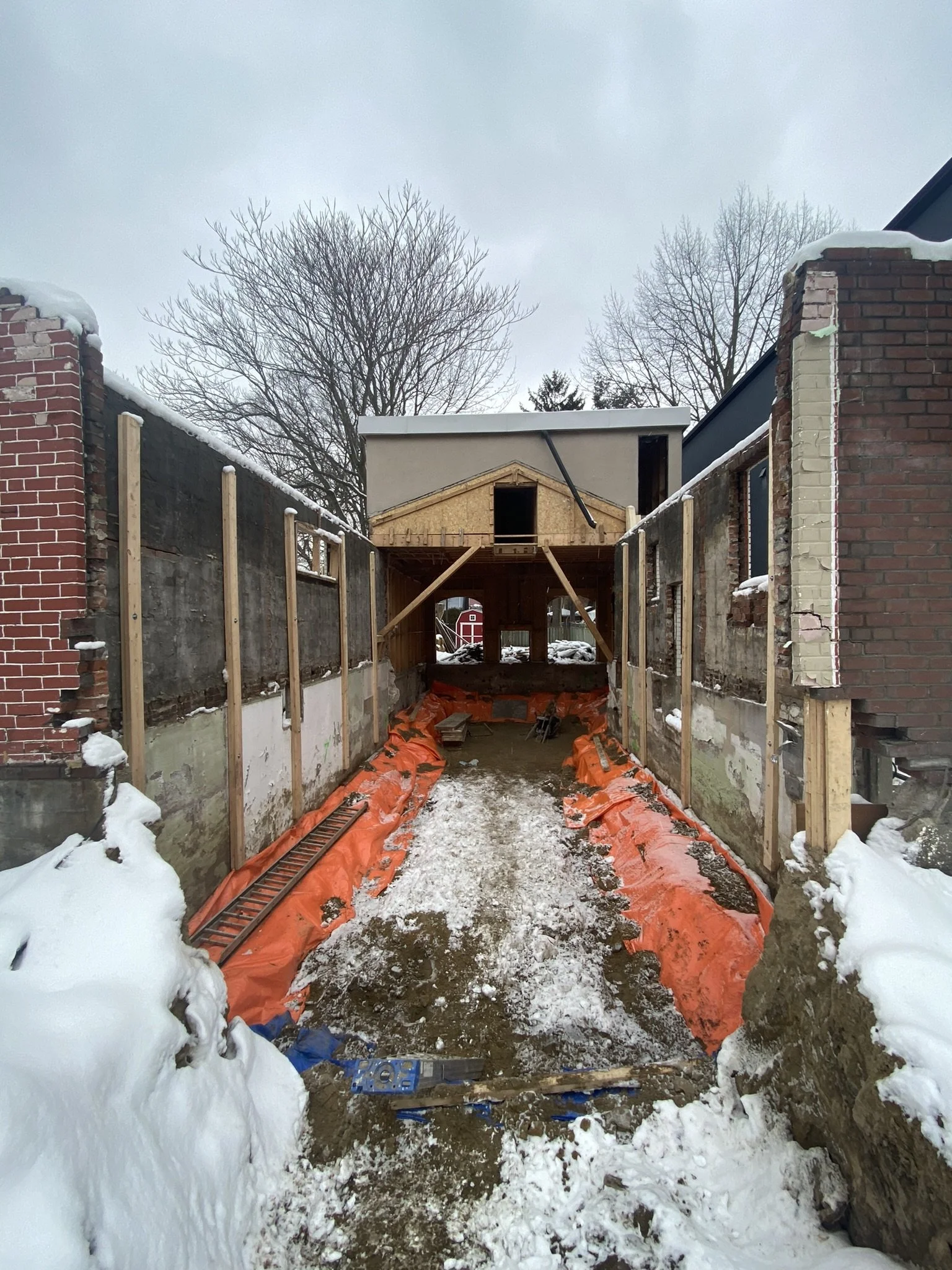 Construction site on a snowy day with an enclosed pathway leading to a raised building under construction, surrounded by brick walls with snow on top.