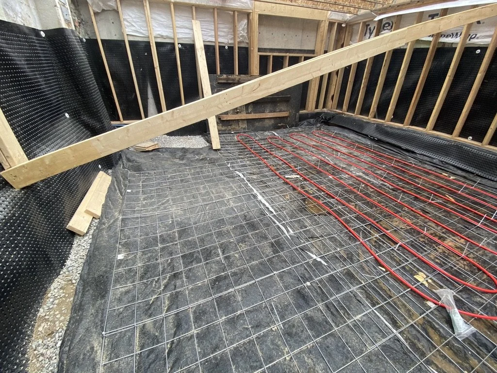 Construction site with a wooden framework, black vapor barrier, red radiant floor heating pipes, and wire mesh on the floor.