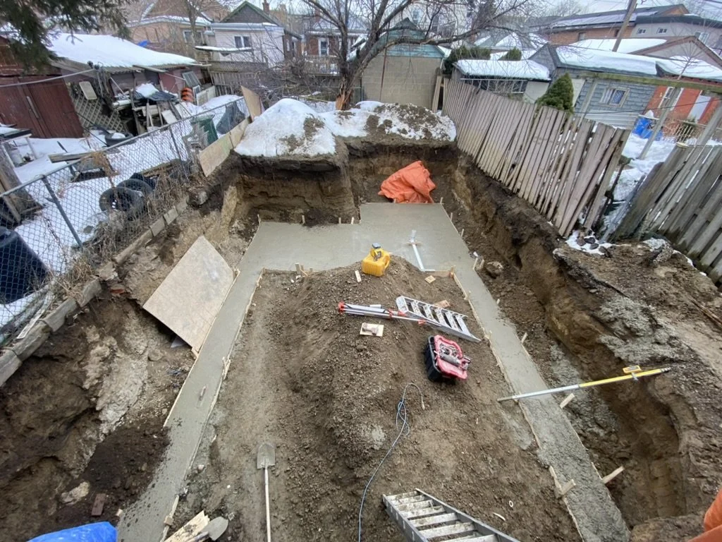 Construction site with freshly poured concrete foundation on a backyard with a dirt excavation, tools, and fencing surrounding the area.