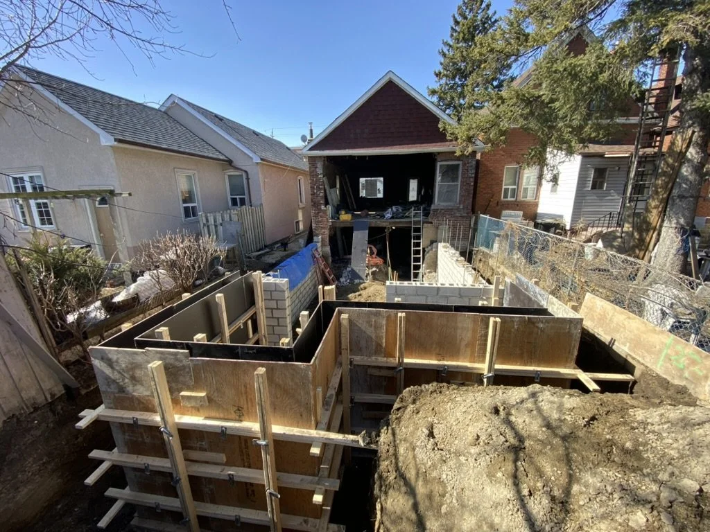 Construction site in a backyard with foundation forms for a new building, neighboring houses, trees, and blue sky.