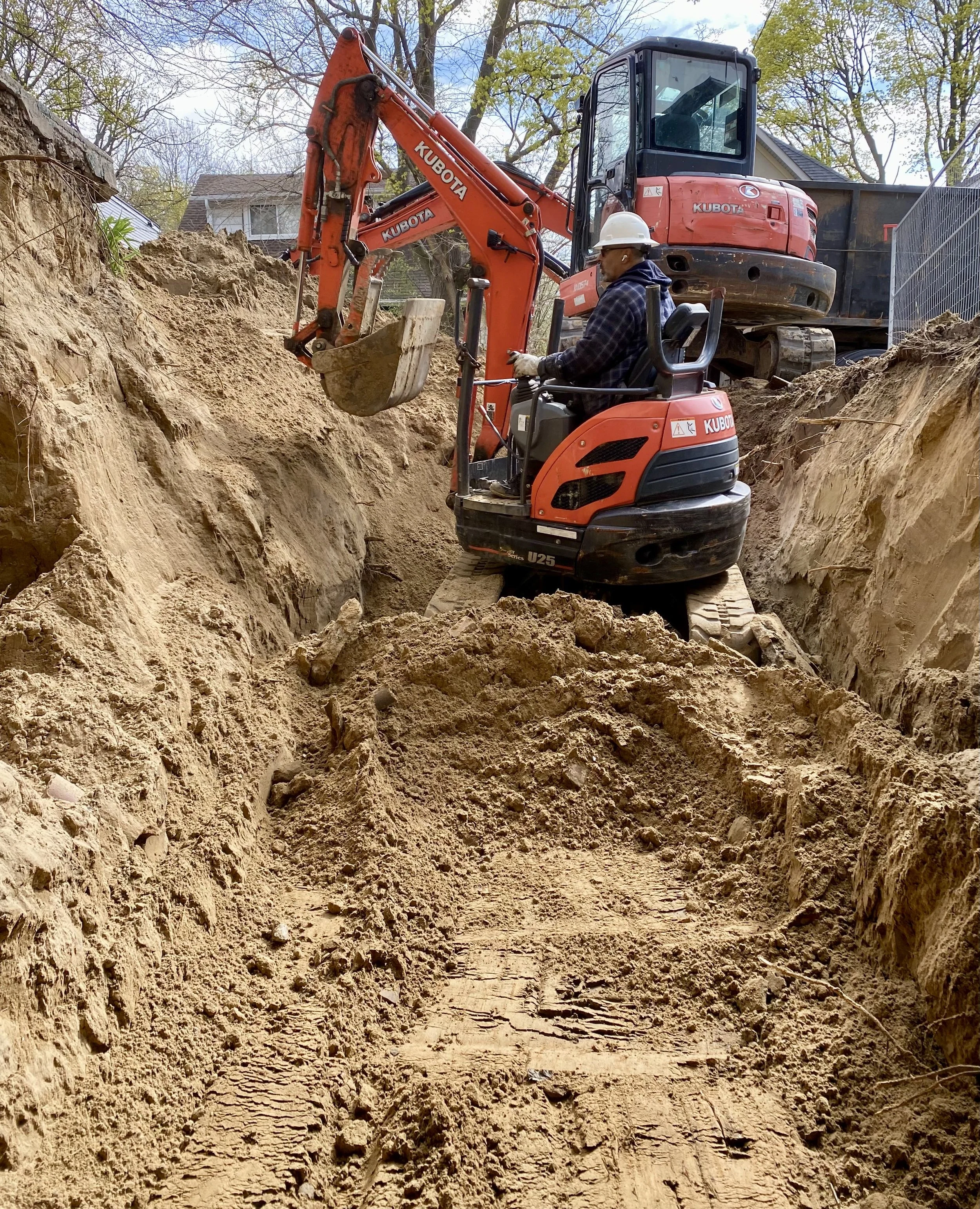 Worker operating a mini excavator to dig a trench in a residential area on a sunny day.