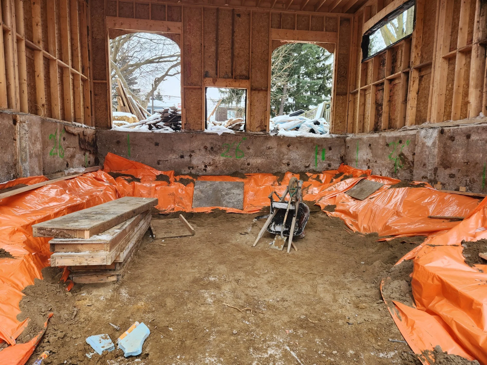 Interior view of a house under construction, with exposed wooden framing, orange waterproofing on the foundation, and construction tools inside, with snow visible through the open windows.
