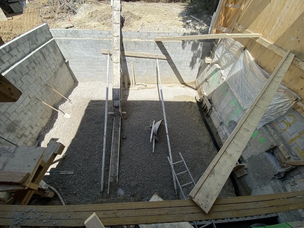 View of a small construction site with gravel on the floor, wooden framing, and cinderblock walls, indicating the early stages of building a structure.