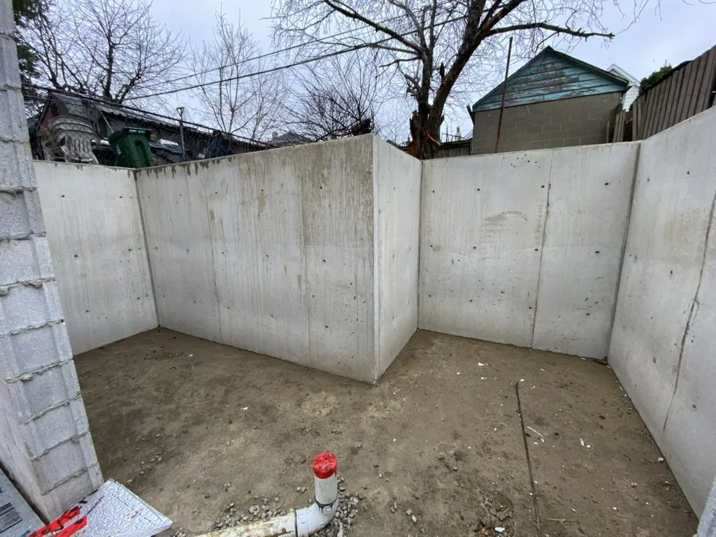 Empty backyard with concrete wall enclosure, dirt ground, and a plastic pipe in the foreground, with leafless trees and houses in the background.