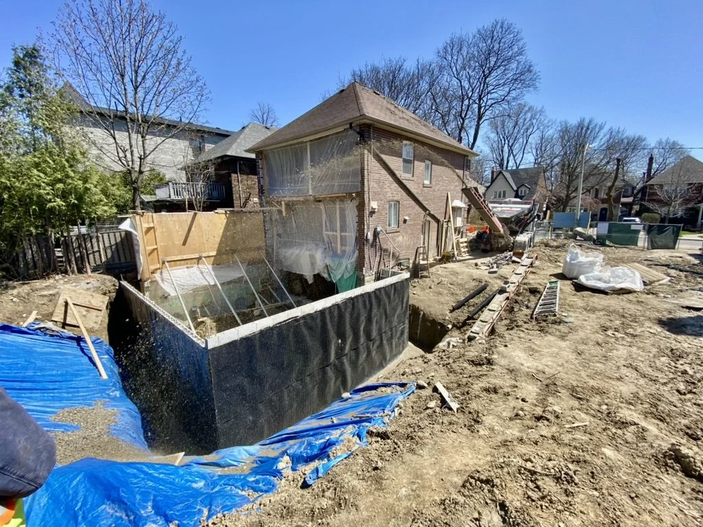 Construction site with excavation and foundation work for a backyard renovation, with a two-story brick house in the background, dirt, construction materials, and tools scattered around.