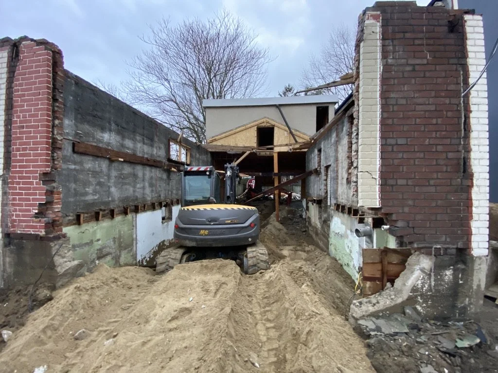 Construction site with a small excavator digging a trench between two old brick walls, with a house under construction in the background.