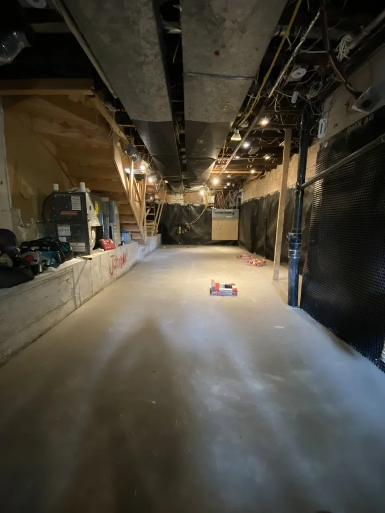 Undeveloped interior space under construction with concrete floor, exposed ceiling and wall wiring, tools, and equipment along the sides.