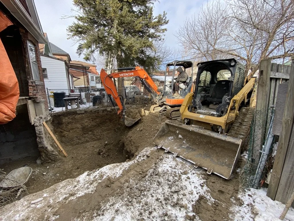 Two construction excavators working on digging a large hole in a backyard, with snow on the ground and residential houses in the background.