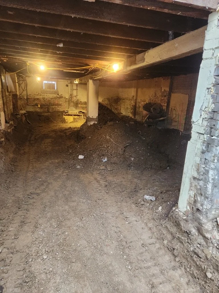 Underground basement or cellar under renovation with dirt floor, exposed brick walls, and construction lighting, including piles of dirt and debris.
