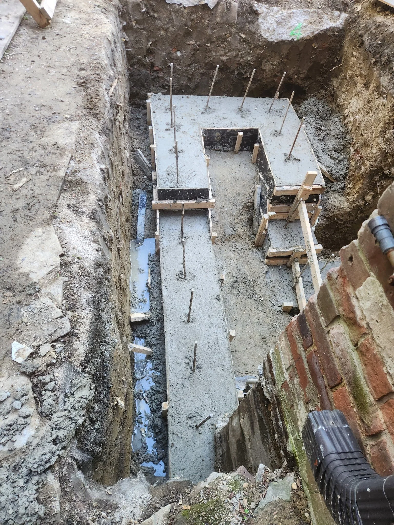 Construction site with freshly poured concrete foundation, wooden formwork, and exposed rebar in a deep trench.
