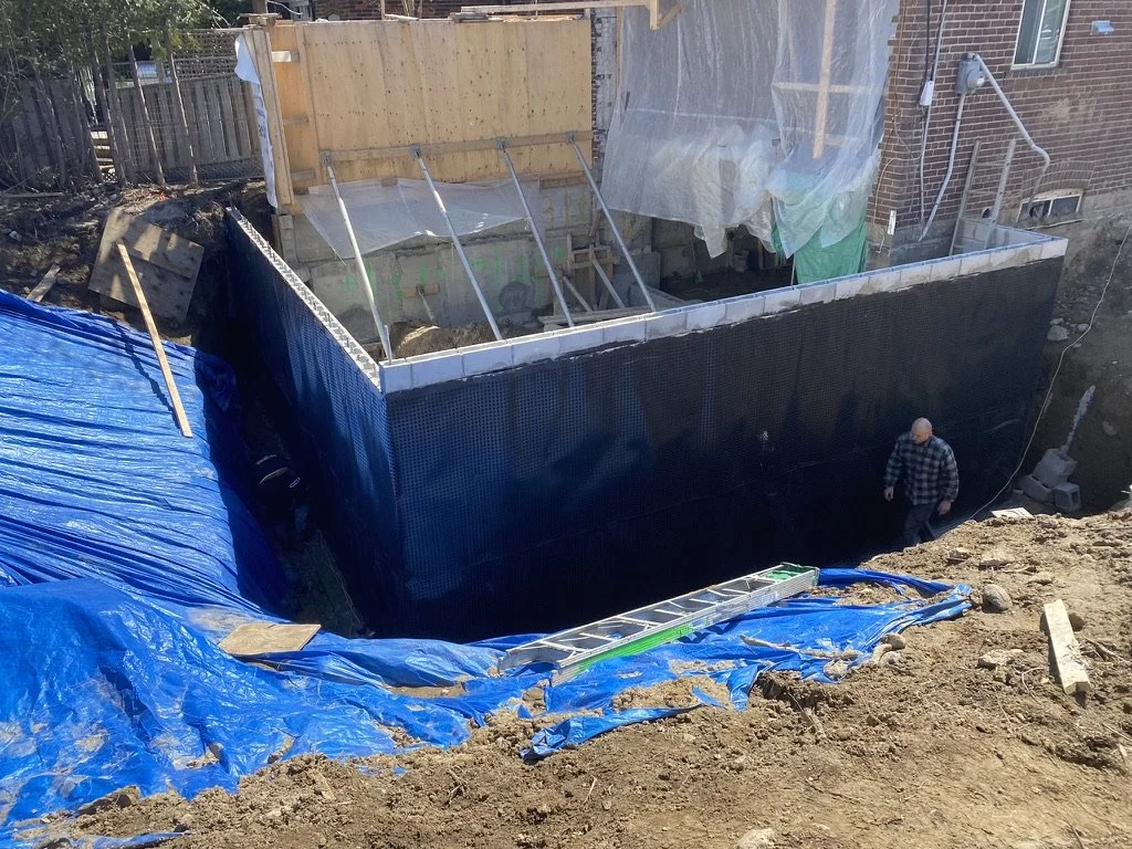 Construction site with a deep excavation and foundation work for a building, with wood, plastic sheeting, and a ladder visible.