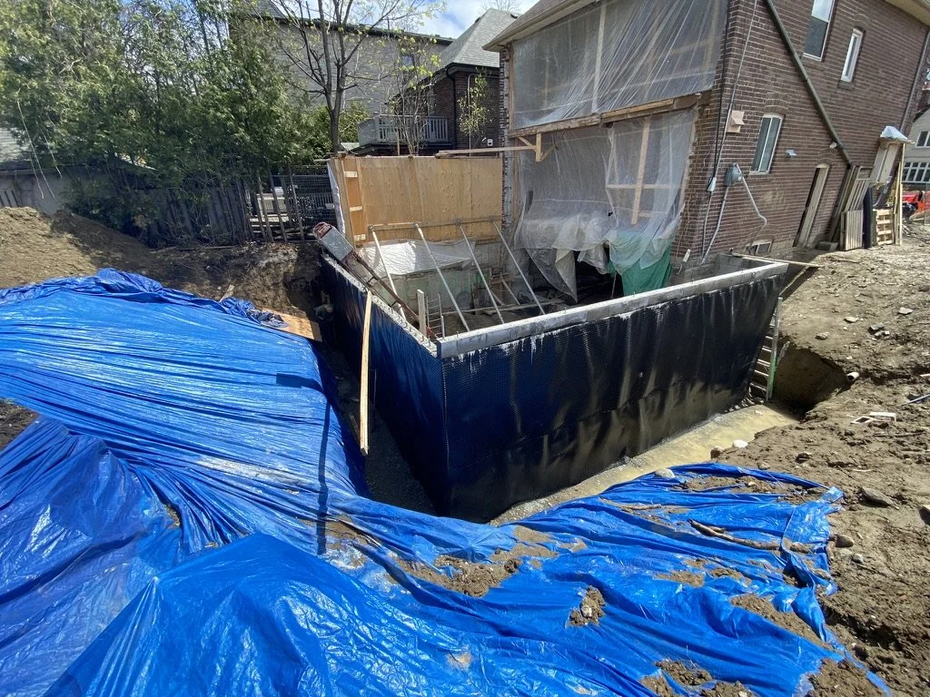 Under construction foundation with blue tarp covered soil, a black exterior wall, and a partially enclosed area with sheer plastic sheeting on a brick house.