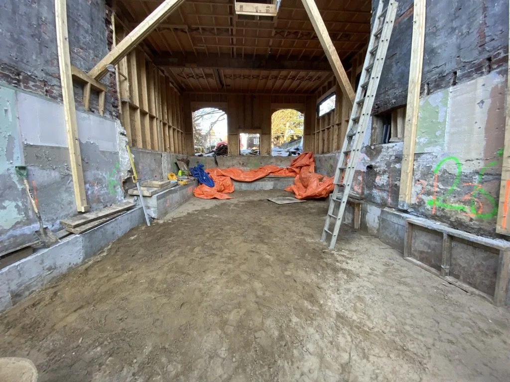 Interior view of a construction site with a wooden frame, ladder, tools, and orange tarp on the dirt floor, looking toward the rear windows of the building.