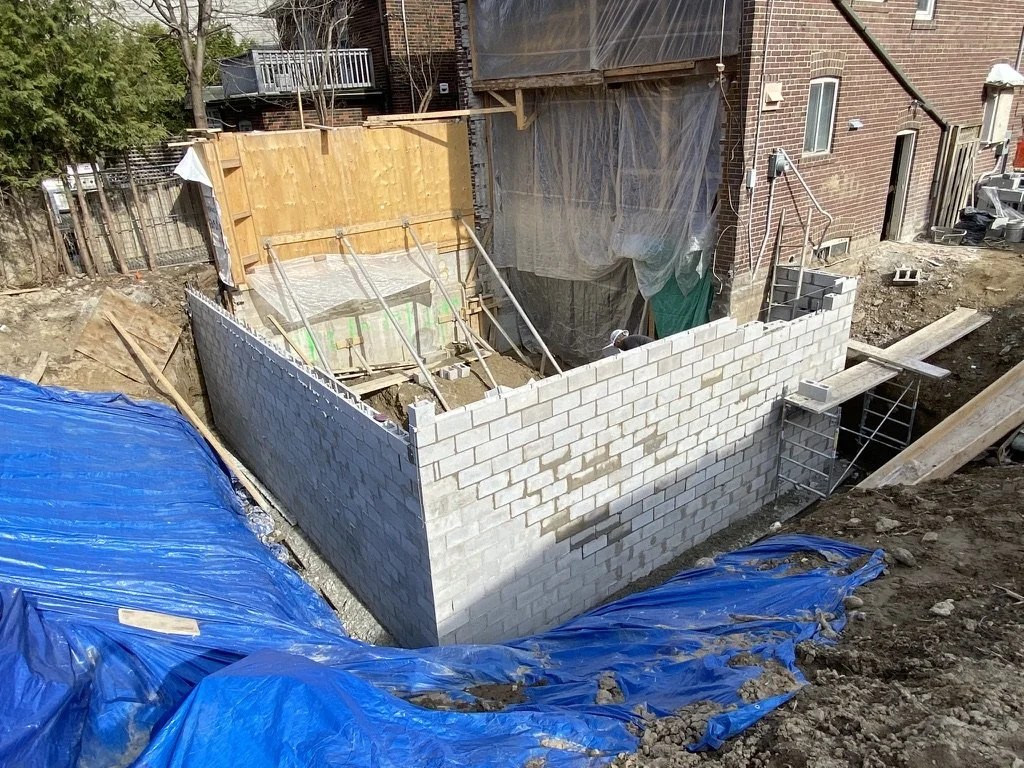 Construction site with partially built cinder block wall, wooden and metal supports, and blue tarps covering the ground.