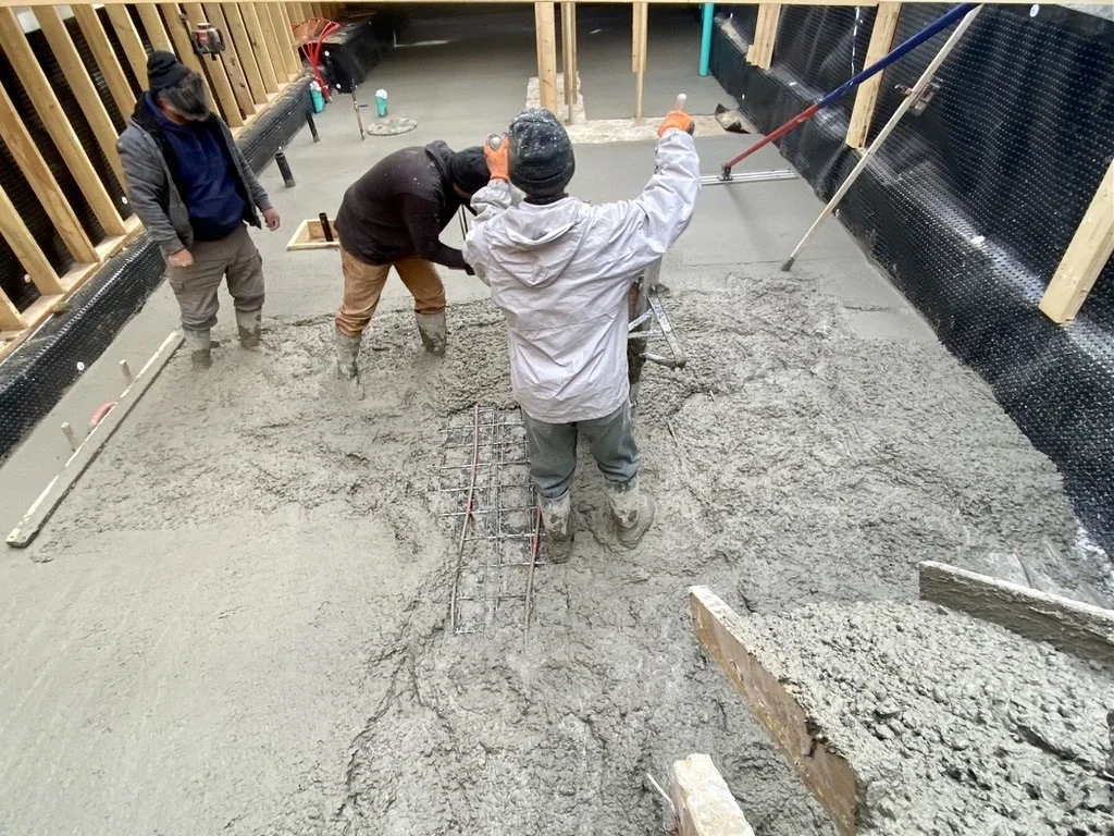Three workers are pouring and spreading concrete to create a floor slab in a construction site, with wooden framing and black waterproofing membrane visible.
