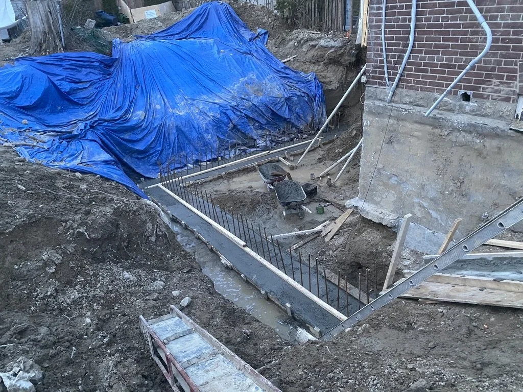 Construction site with a foundation being prepared, exposed rebar, a blue tarp covering part of the area, and various construction tools and materials around.