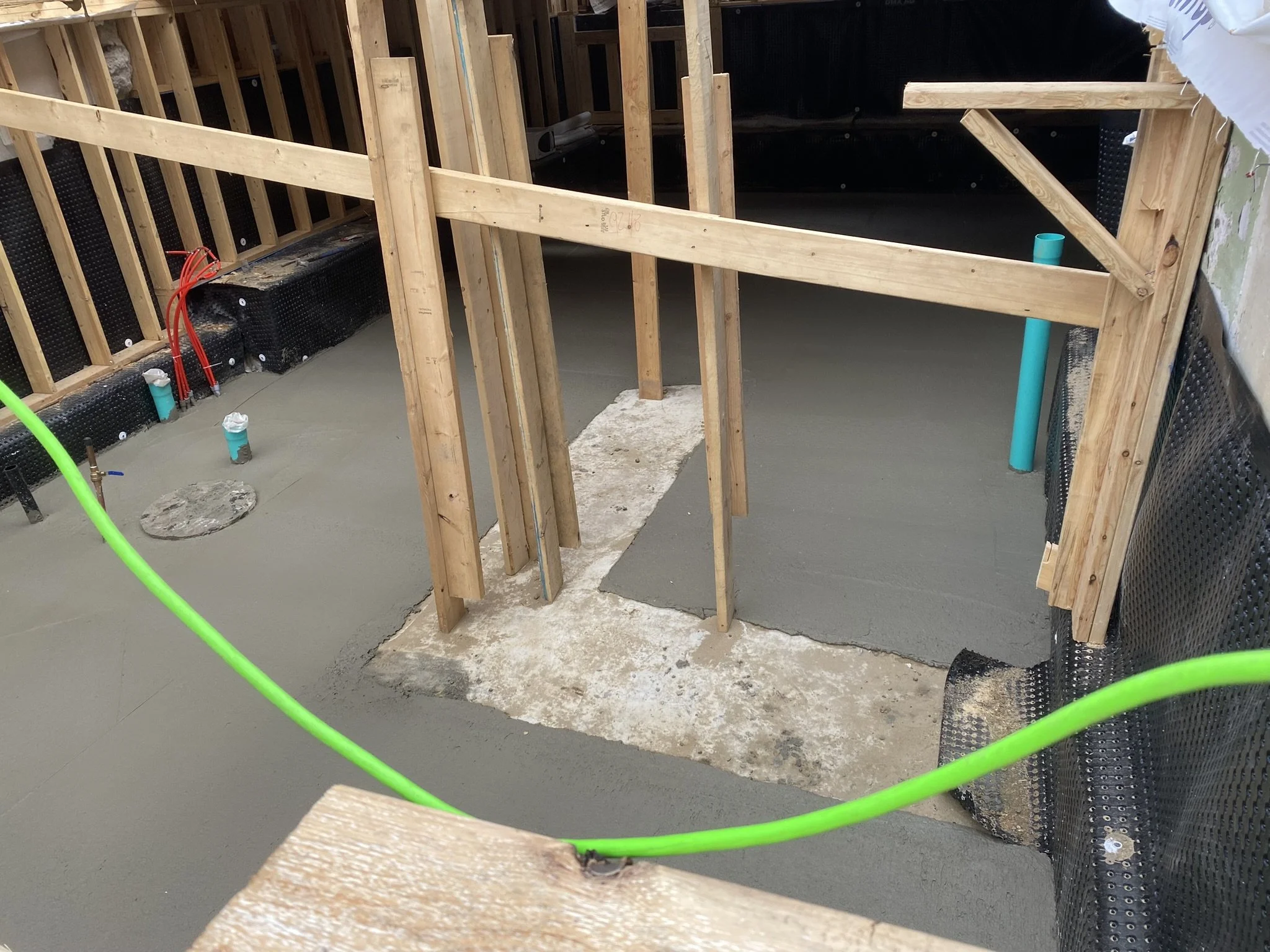 Construction site showing a poured concrete foundation with wooden framing and plumbing pipes.