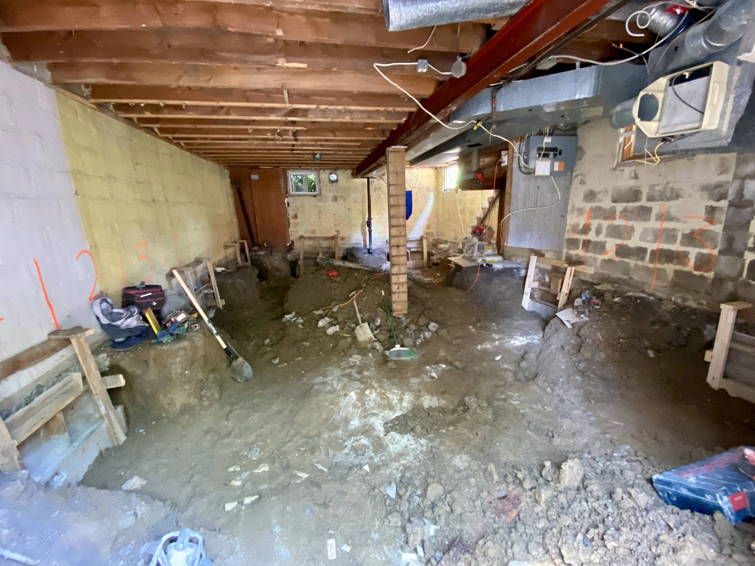 Interior of a basement under renovation, with exposed wooden ceiling beams, dirt floor, and construction tools and supplies scattered around. Walls have markings and wood framing for future work.